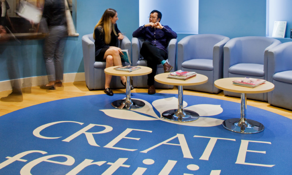 Two people sit and converse in a waiting area with blue walls and chairs. The floor features a large blue circle with the text 