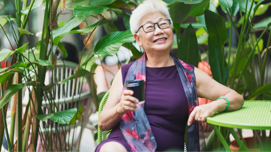 Elderly woman with short white hair and glasses, wearing a maroon dress and scarf, sits outdoors in a garden setting, smiling and holding a black coffee cup. She’s discussing the benefits of pension schemes for women with her friends.