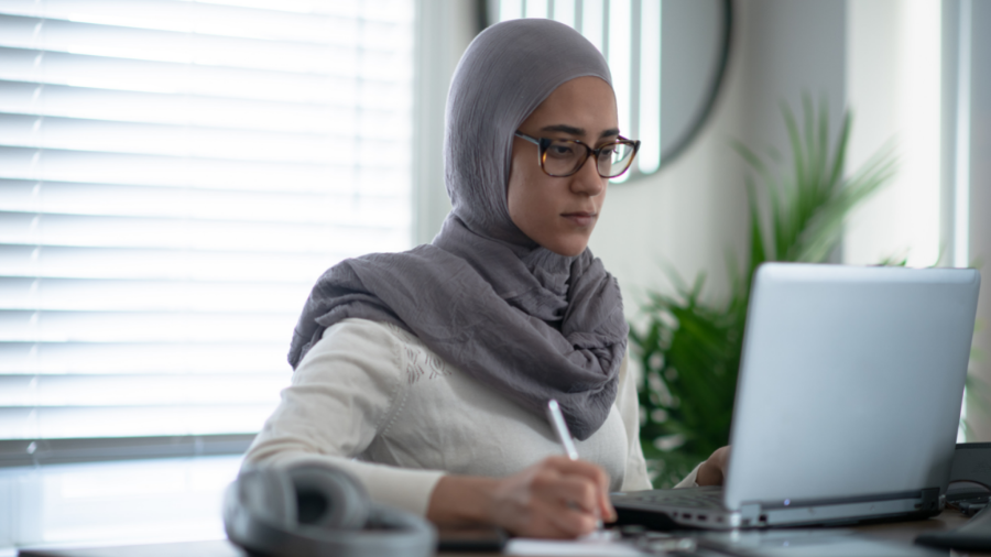 A person wearing glasses and a headscarf works on a laptop, demonstrating their managers skills, holding a pen in one hand. The background includes a plant and a window with blinds.