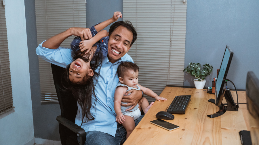A man smiles while holding a laughing child and a baby at a desk with a computer and phone, embracing the joy of remote work during the Omicron phase before the return to office.