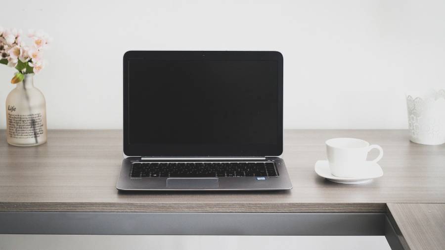 A laptop with a black screen sits on a wooden desk, ready for the day's tasks in Human Resources. To its left is a vase with flowers, adding a touch of nature to the workspace, and to its right is a white cup and saucer, perfect for those personnel meetings.