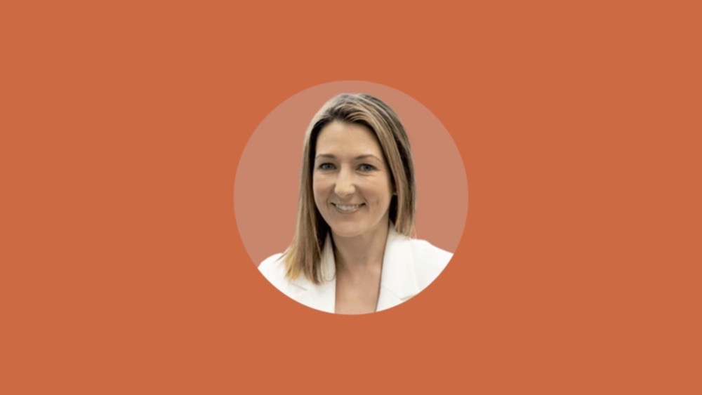 Amy Crawford, with her shoulder-length blonde hair, is smiling and wearing a white blazer against an orange background. Known for advocating diversity in recruitment, she continues to inspire UK employers with her efforts.