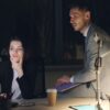 A man sits on a desk while a woman beside him looks at a monitor, in a night setting at an office.