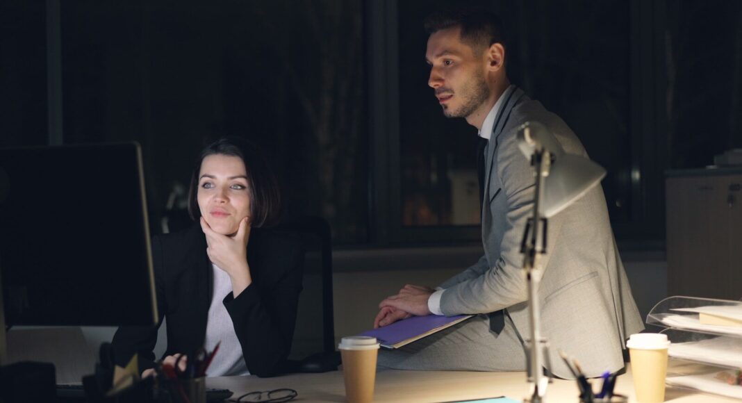 A man sits on a desk while a woman beside him looks at a monitor, in a night setting at an office.