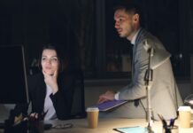 Low harassment reporting may hide workplace misconduct, employers warned A man sits on a desk while a woman beside him looks at a monitor, in a night setting at an office.