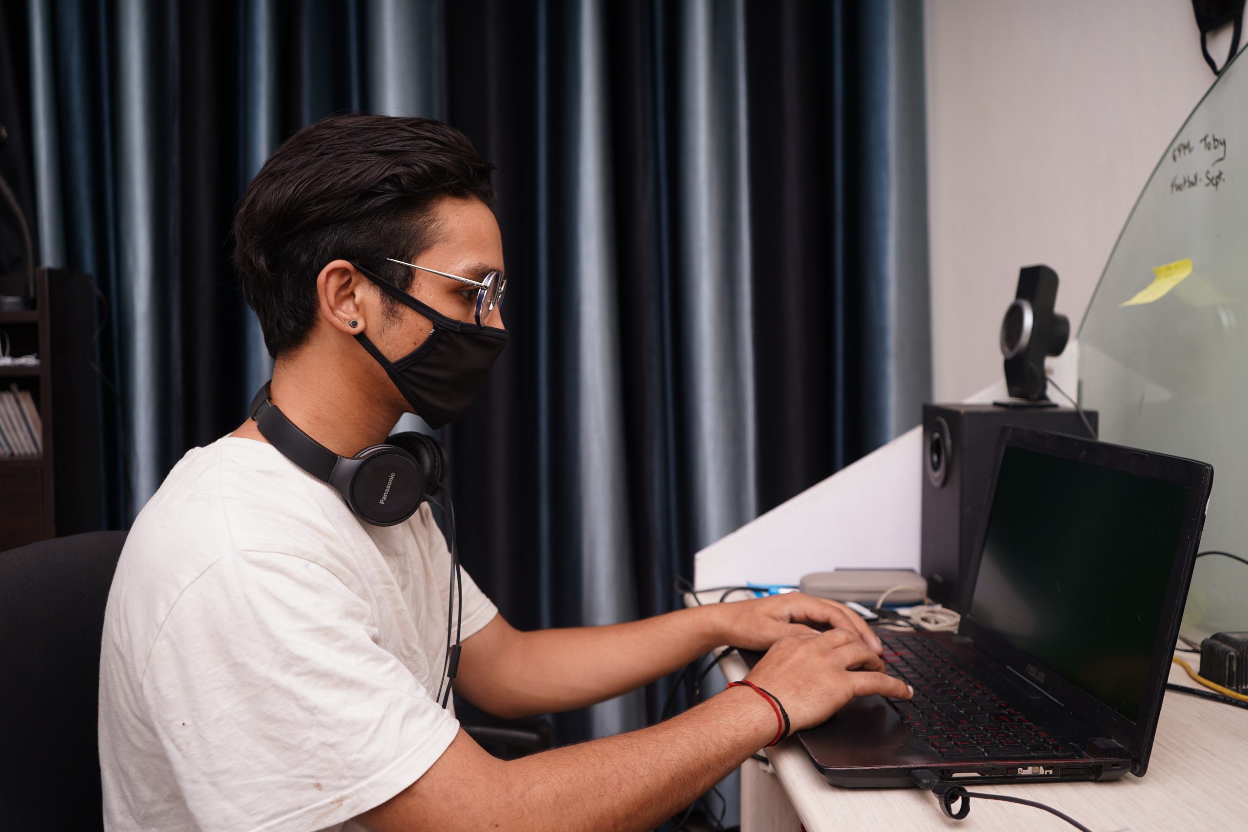 A person wearing a mask and headphones is typing on a laptop at a desk, which is adorned with a monitor, speakers, and paperwork about their workplace pension.