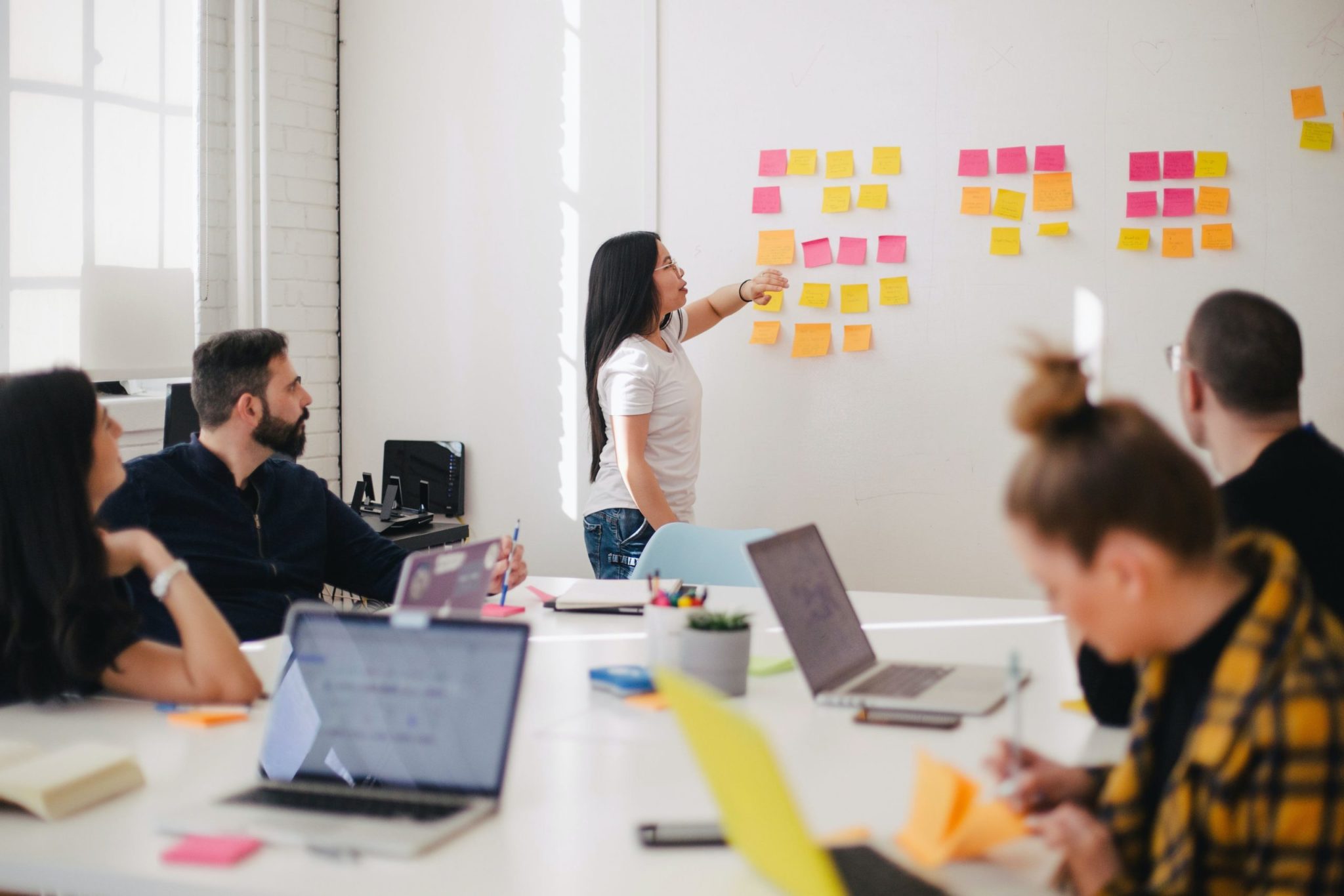 A group of five people in a meeting room. One person from Human Resources is standing and pointing at sticky notes on a whiteboard while the others are seated at a table with laptops and notebooks.