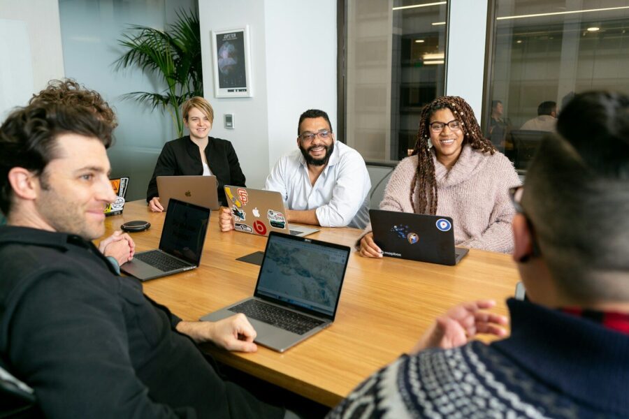 Four people all on laptops, two men and two women, listen to person talking in a board meeting